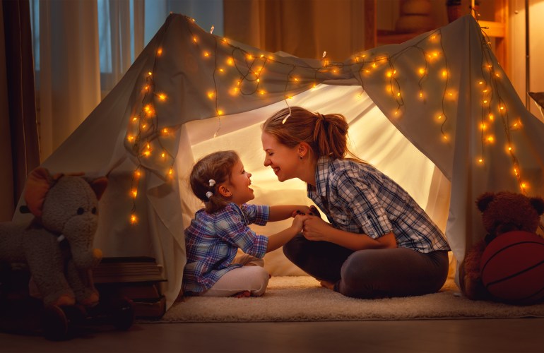 A mother and daughter laugh and enjoy each other in a tent made of sheets and twinkle lights